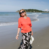 Woman in coral shirt and black floral pants standing on a beach with ocean and sky in the background
