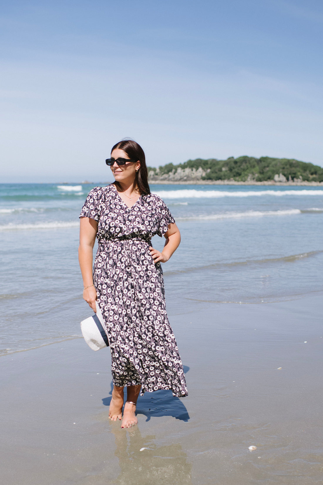 Woman in a floral dress standing on a beach with ocean waves and sky in the background