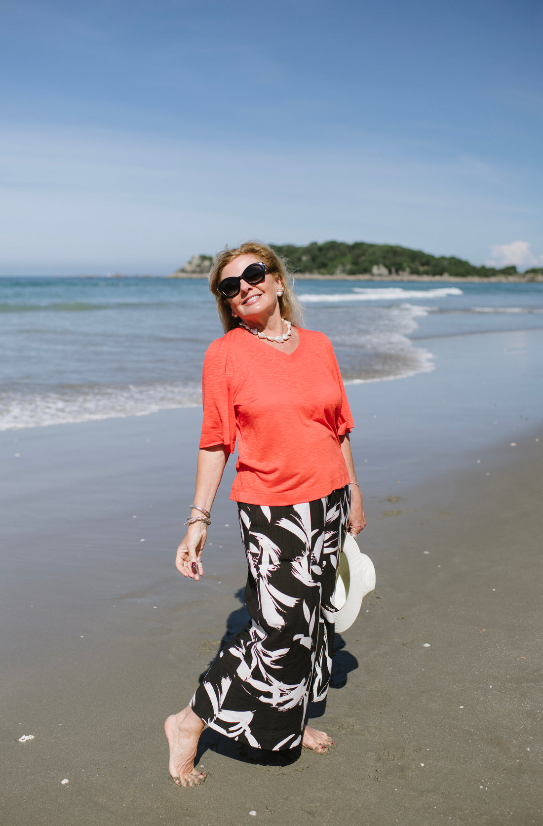 Woman in coral shirt and black floral pants standing on a beach with ocean and sky in the background