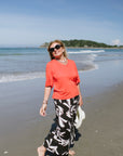 Woman in coral shirt and black floral pants standing on a beach with ocean and sky in the background